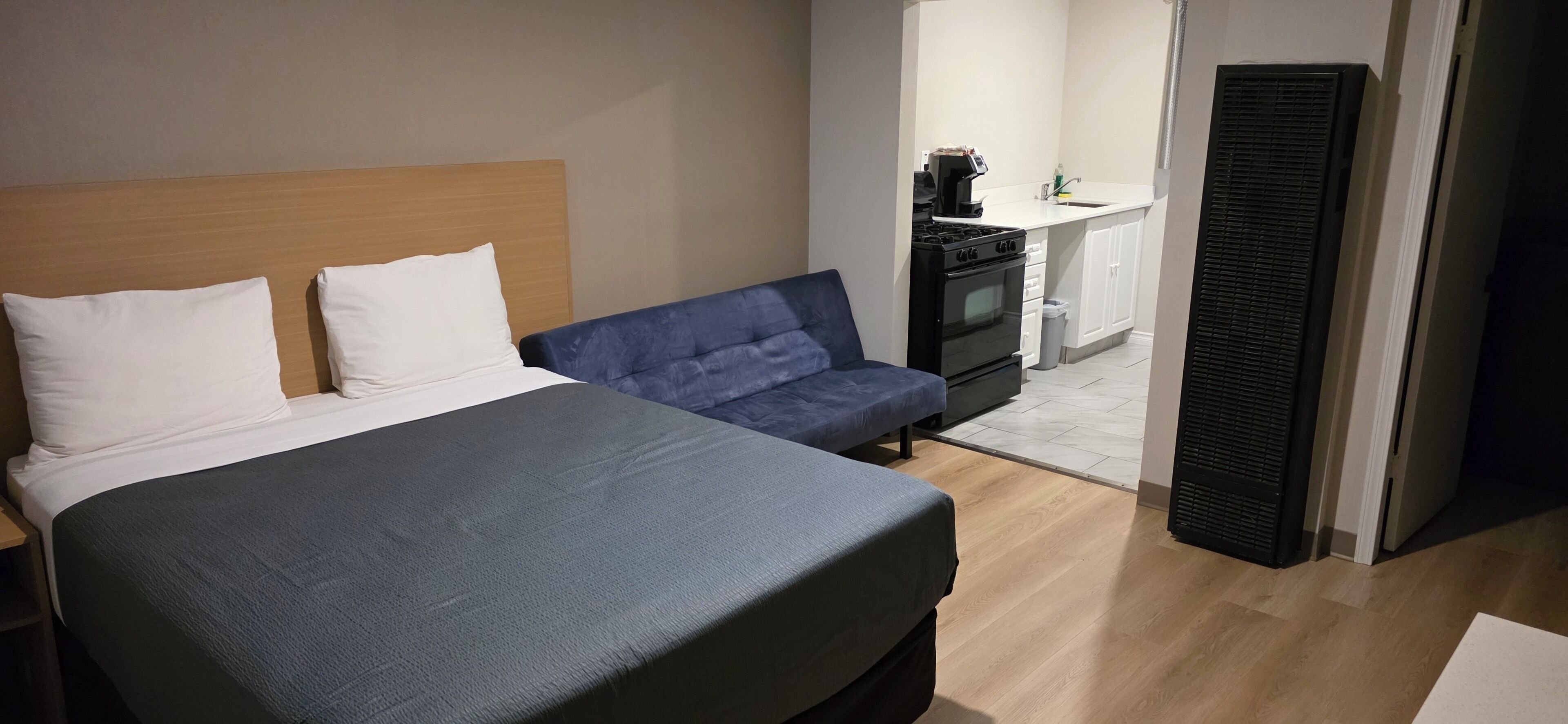 Un dormitorio con una cama, un sofá azul, una kitchenette con estufa y lavaplatos, y una puerta con ventila.