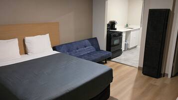 Une chambre à coucher comprenant un lit, un canapé bleu, une kitchenette avec une cuisinière et un évier, et une porte dotée d’une grille d’aération.