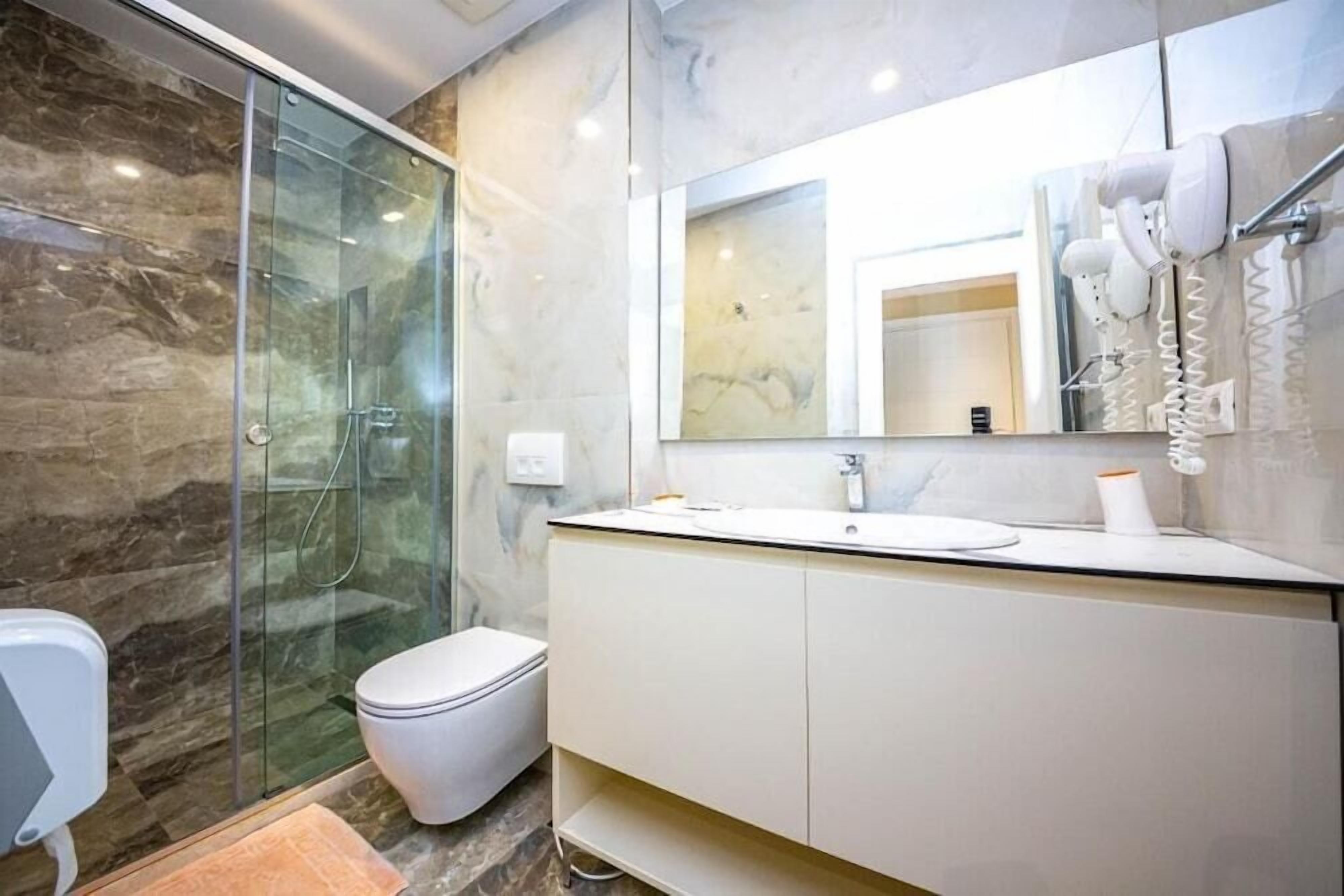A bathroom with a glass-enclosed shower, a toilet, a sink with a mirror above, and a hairdryer mounted on the wall.