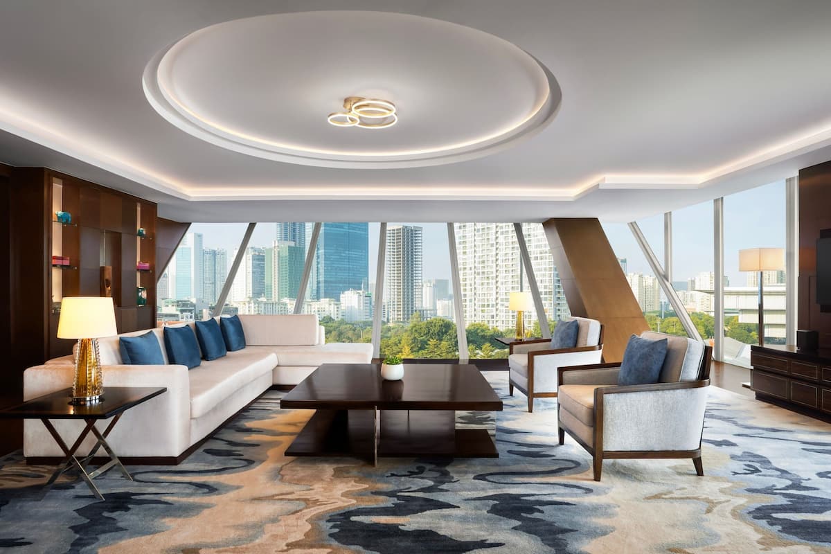 Quarto de hotel moderno com um amplo sofá em forma de L, uma mesa de centro e vista para o horizonte da cidade através de janelas do chão ao teto.