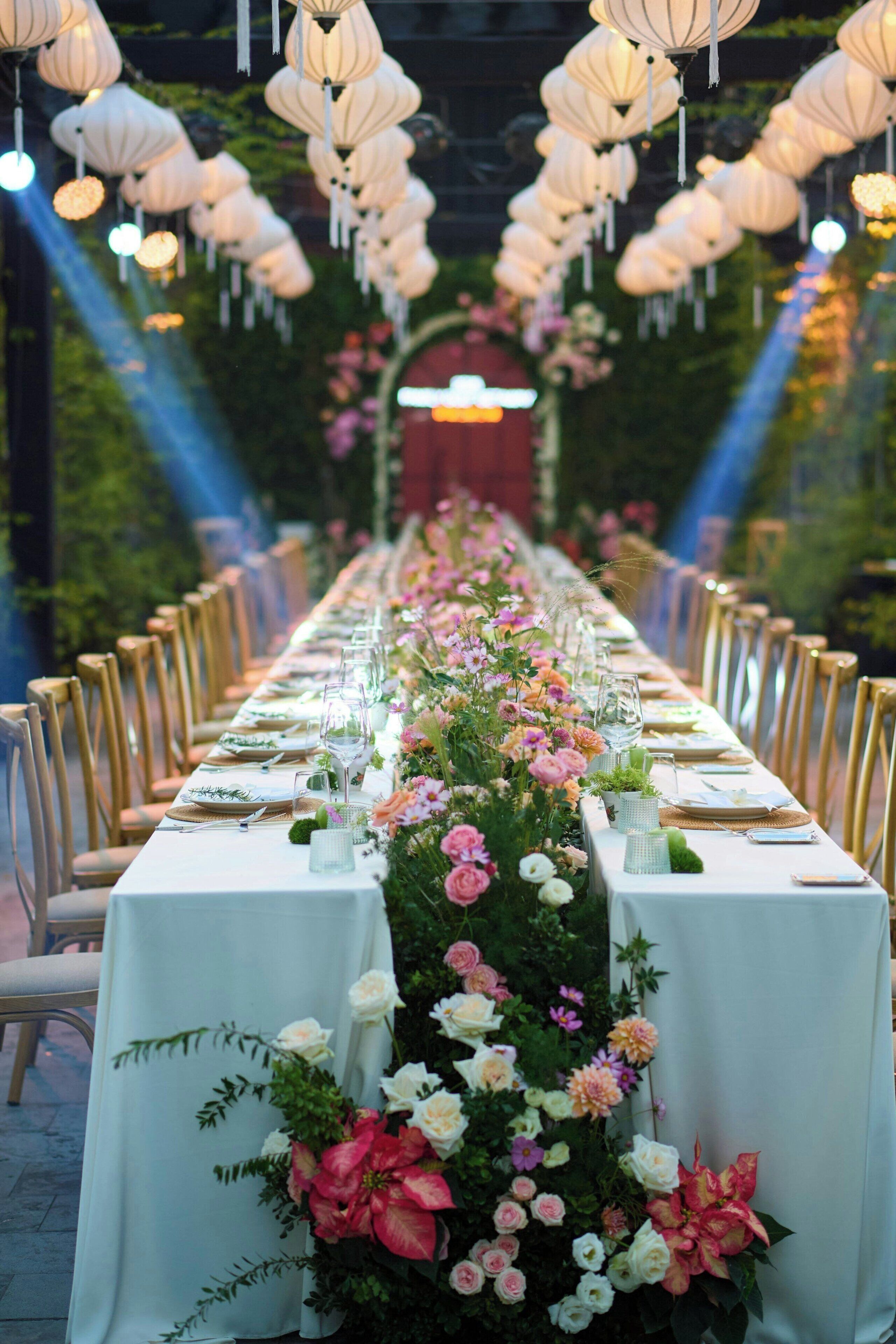 A long dining table set with chairs, surrounded by floral arrangements and hanging lanterns.