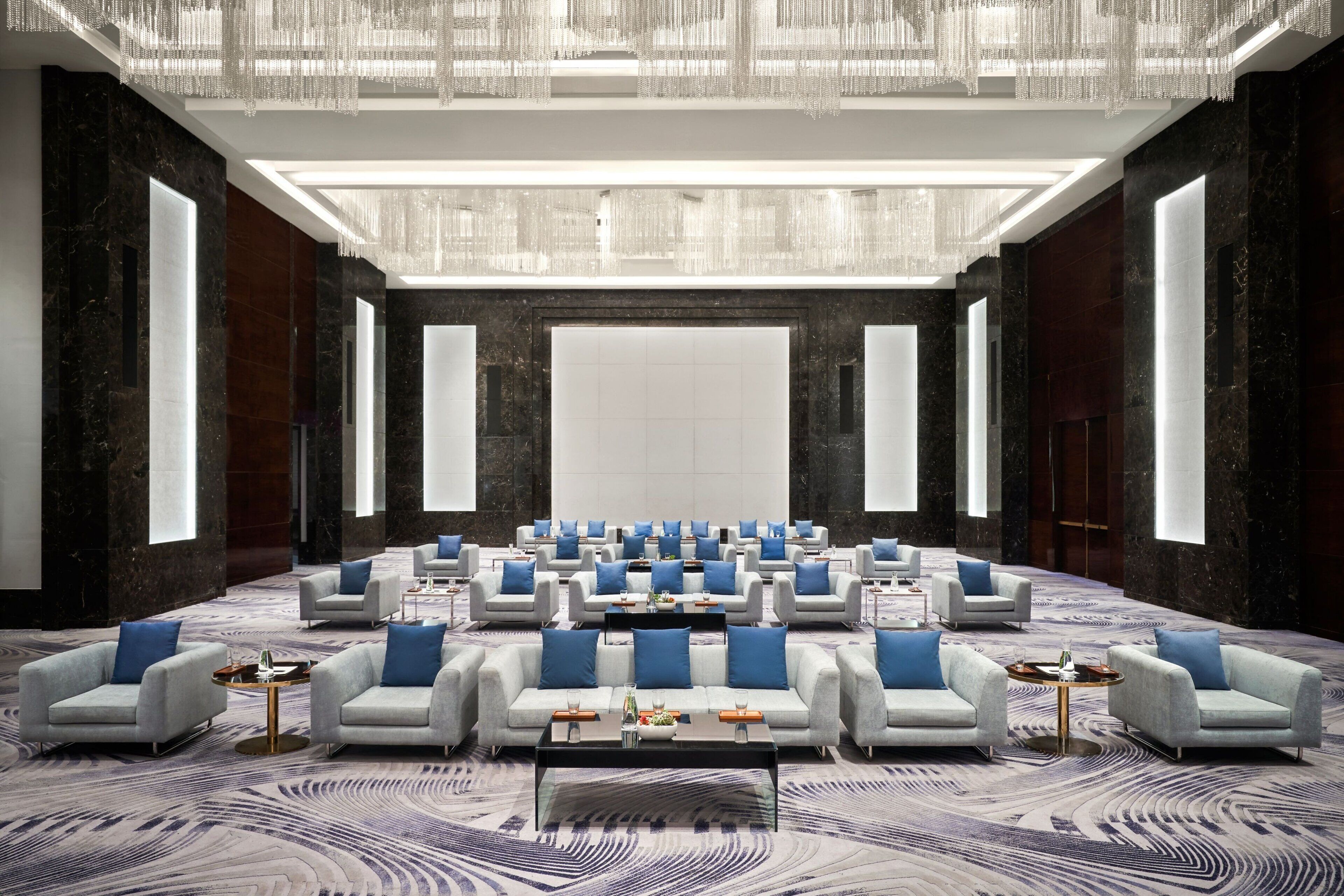 Une salle de conférence spacieuse, avec des rangées de canapés blancs et des coussins bleus, une moquette à motifs et un lustre moderne.