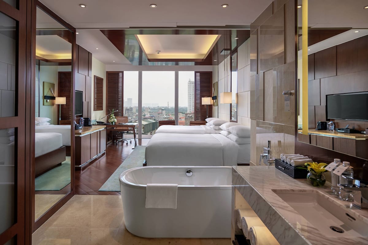 Quarto de hotel moderno com uma banheira grande, vista para a cidade e um quarto separado com cama e escrivaninha.