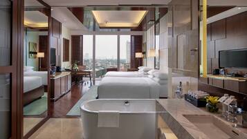 A modern hotel room with a large bathtub, a city view, and a separate bedroom with a bed and a desk.
