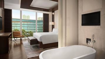 A modern hotel room with a large bed, a sitting area, a bathtub, and a TV mounted on the wall.