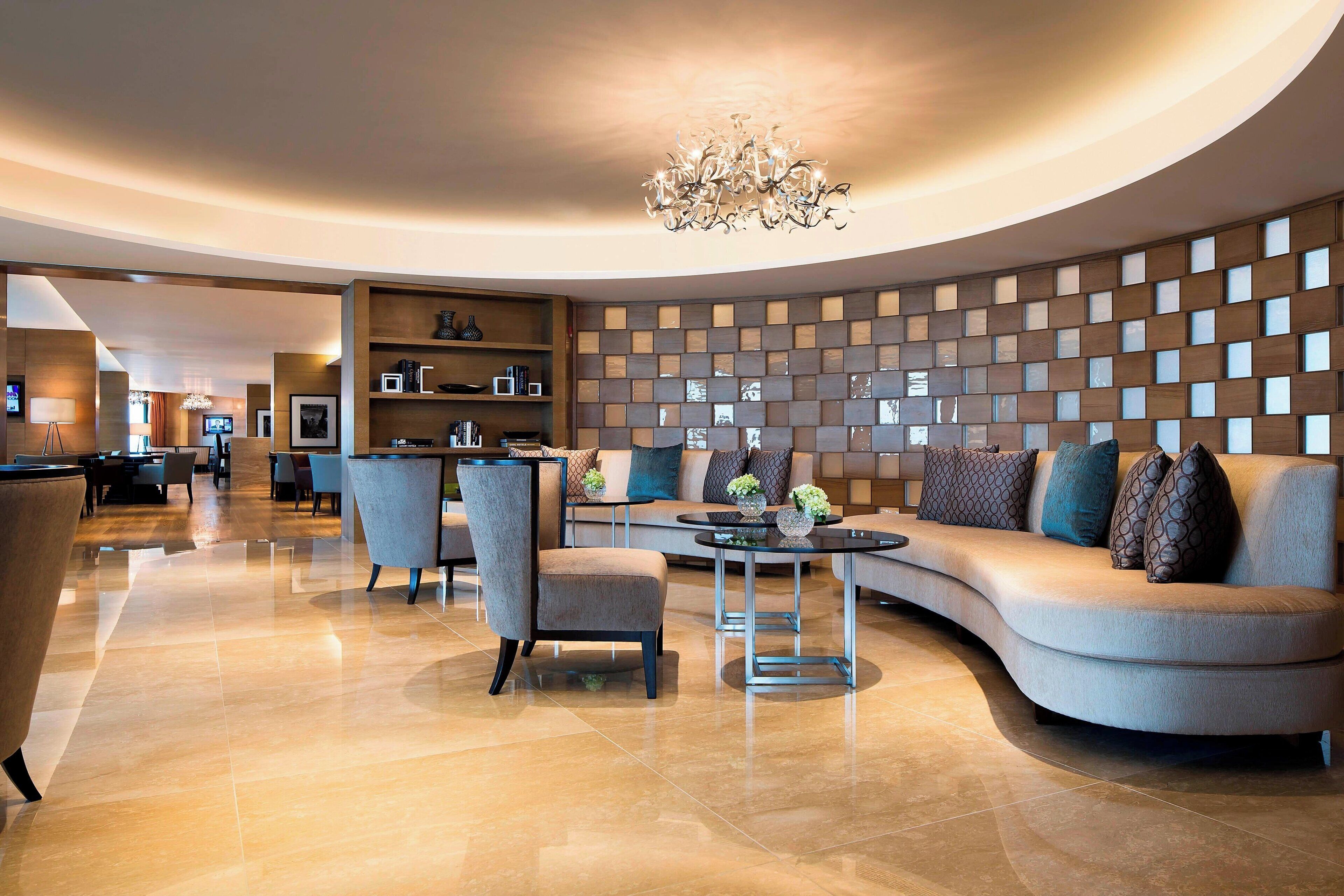 A modern living room with a curved sofa, armchairs, a coffee table, and a chandelier. There is a wall with a geometric pattern and a television in the background.