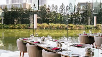 A dining area with tables set for a meal, overlooking a body of water and a garden with trees.