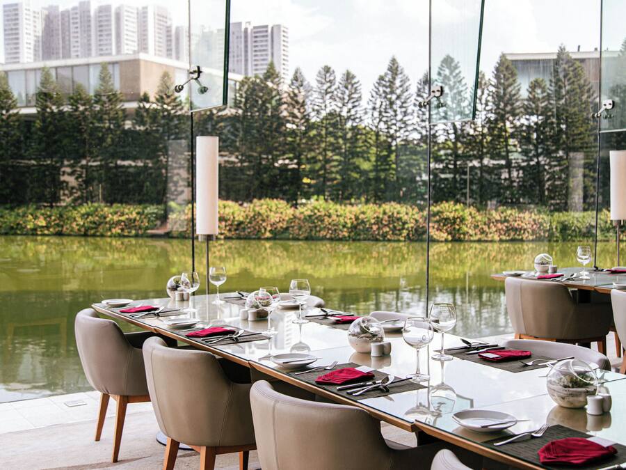 A dining area with tables set for a meal, overlooking a body of water and a garden with trees.