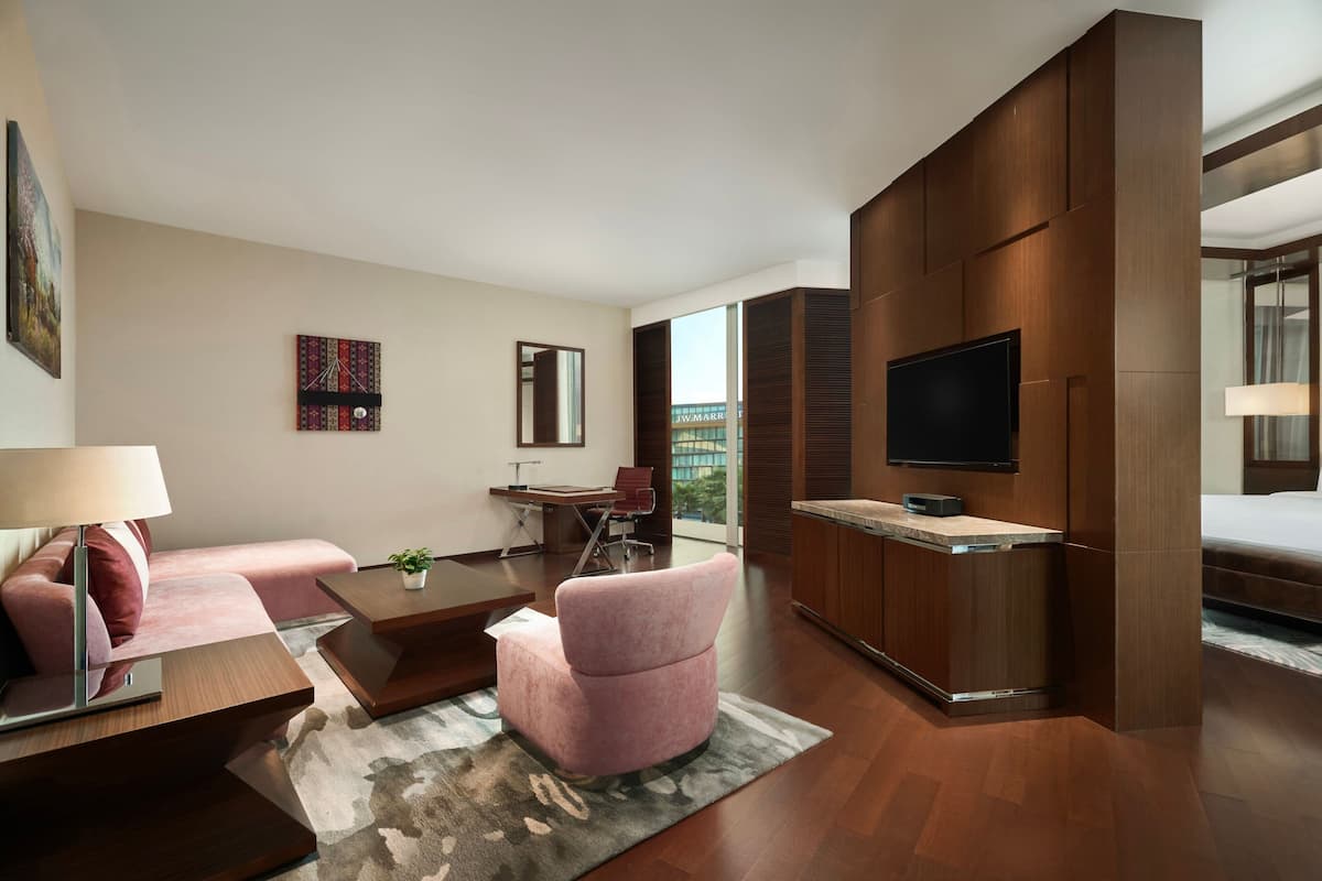 Quarto de hotel moderno com piso de madeira, sofá, escrivaninha, TV fixada na parede e uma grande janela com vista para a cidade.