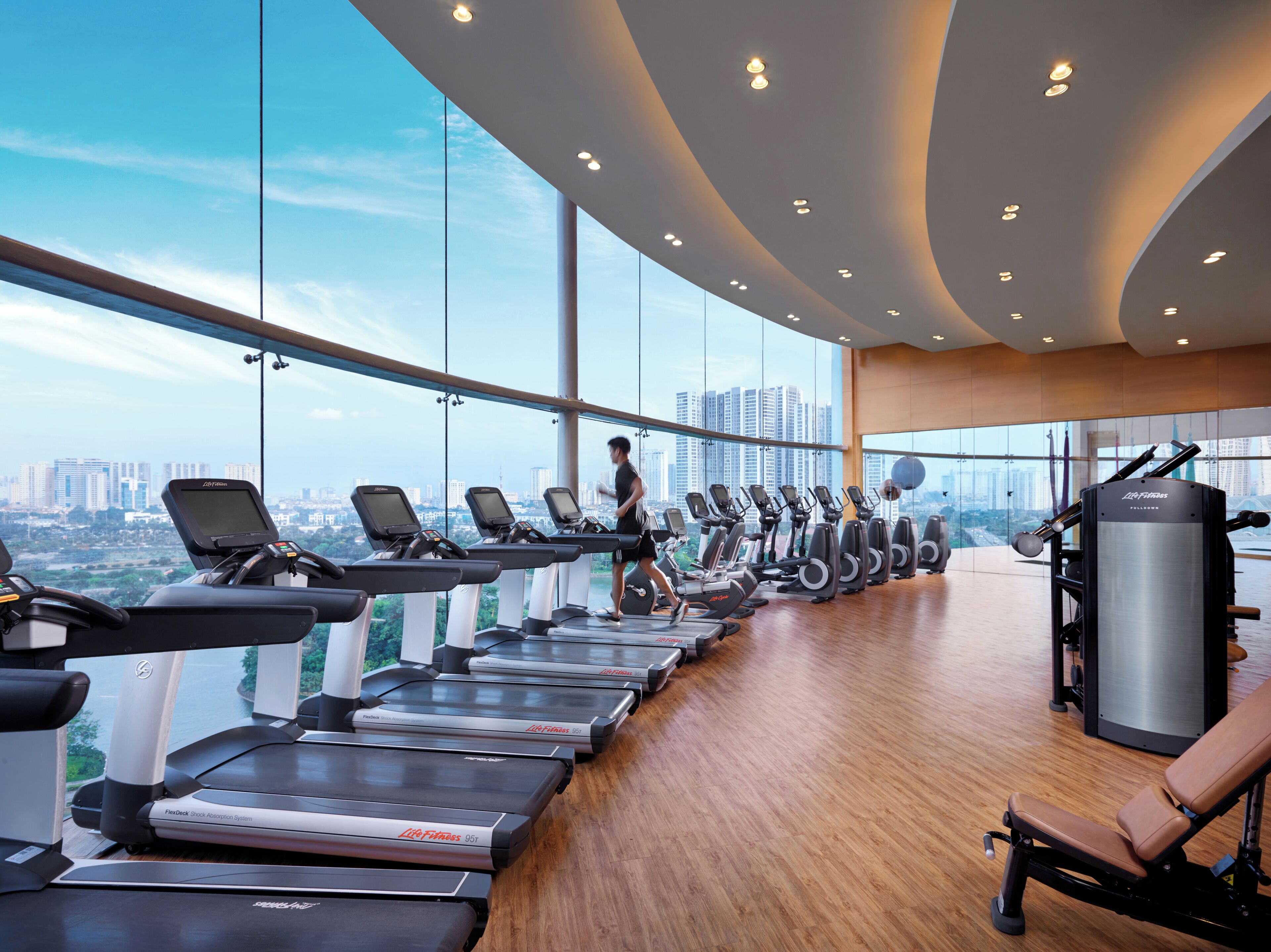 Ein Fitnessstudio mit Laufbändern, Crosstrainern und Kraftgeräten, mit Blick auf die Stadt durch große Fenster.