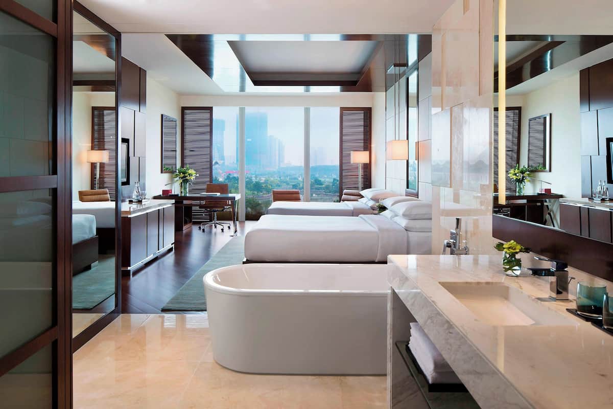 Quarto de hotel moderno com banheira grande, cama de casal, escrivaninha e vista para a paisagem urbana.