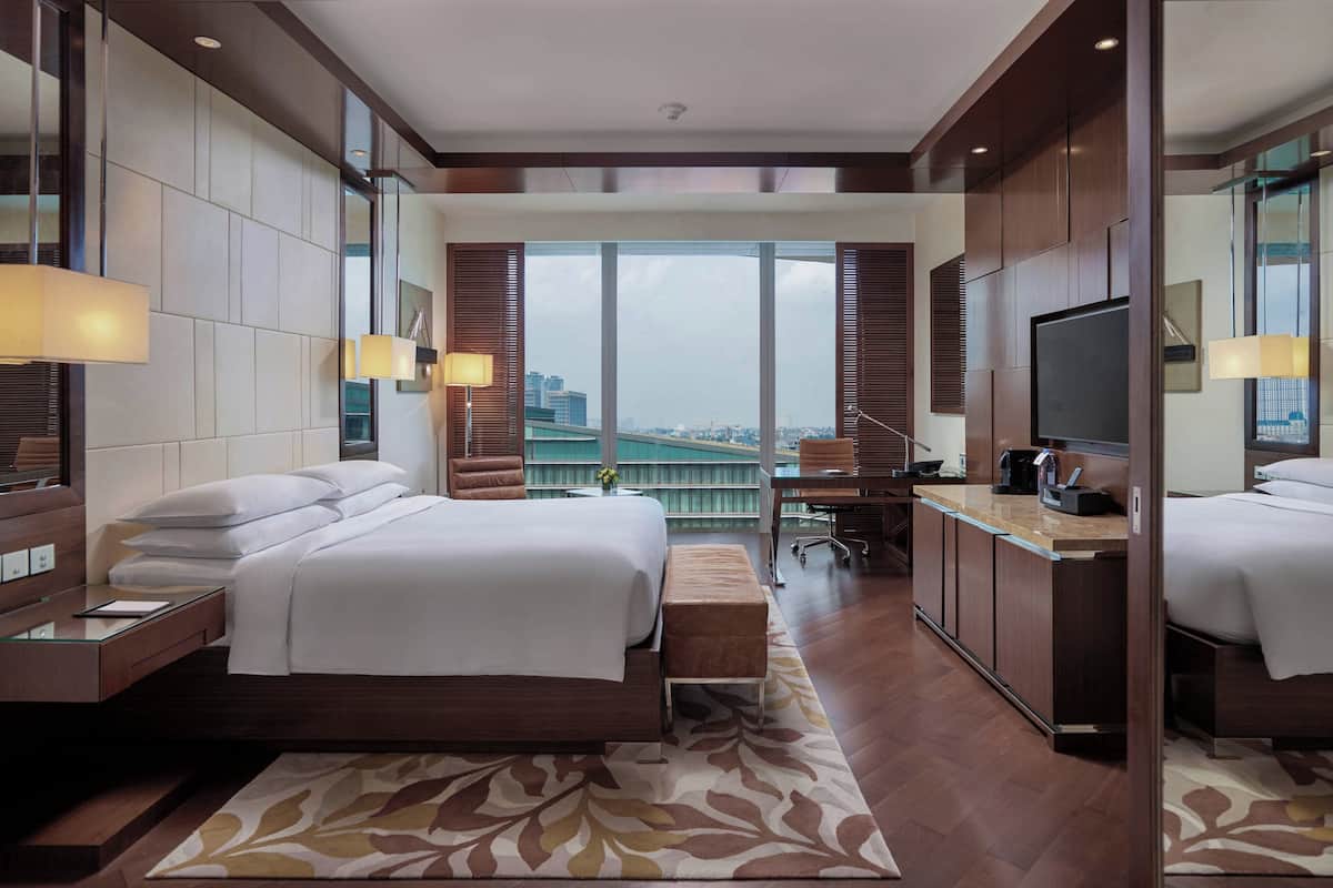 Quarto de hotel moderno com uma cama grande, uma mesa, uma televisão e vista para a cidade.