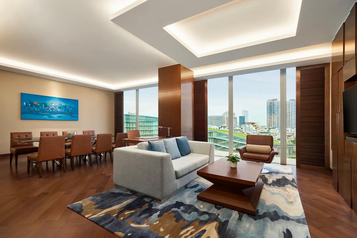 Quarto de hotel moderno com área de jantar, sofá, mesa de centro e grandes janelas com vista para a cidade.