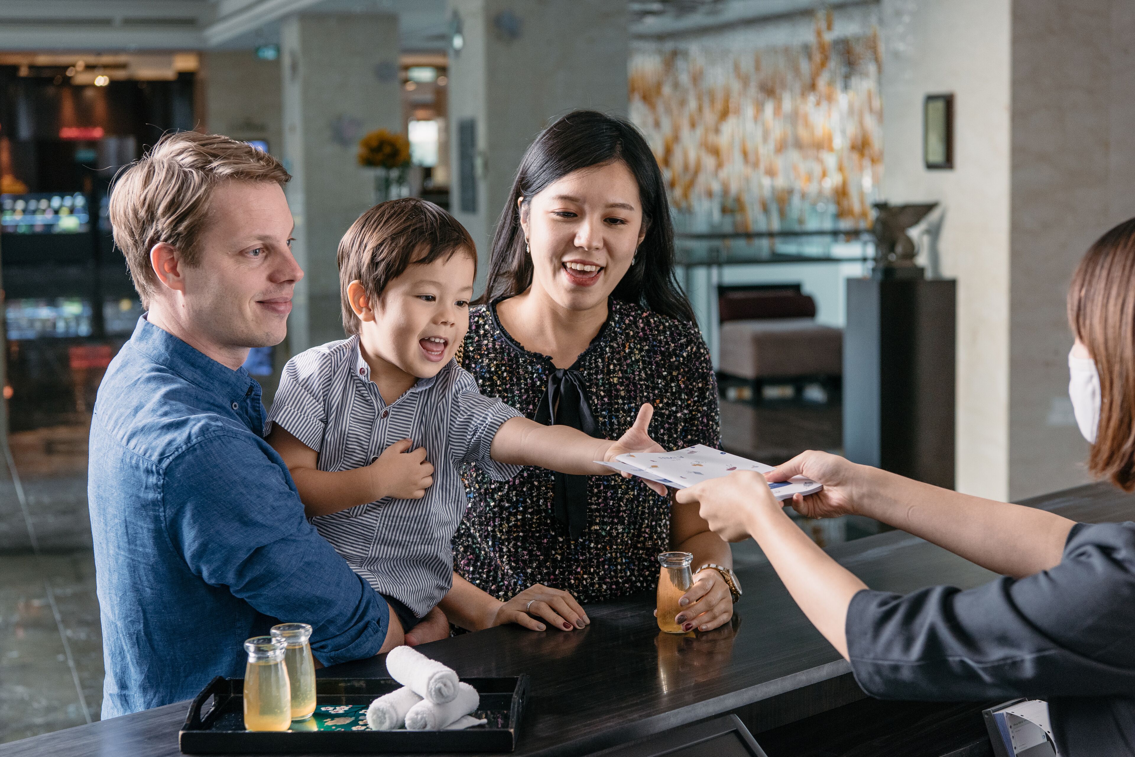 A hotel reception area with a counter, a customer interacting with a staff member, and a family with a child.
