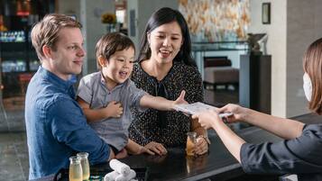 A hotel reception area with a counter, a customer interacting with a staff member, and a family with a child.