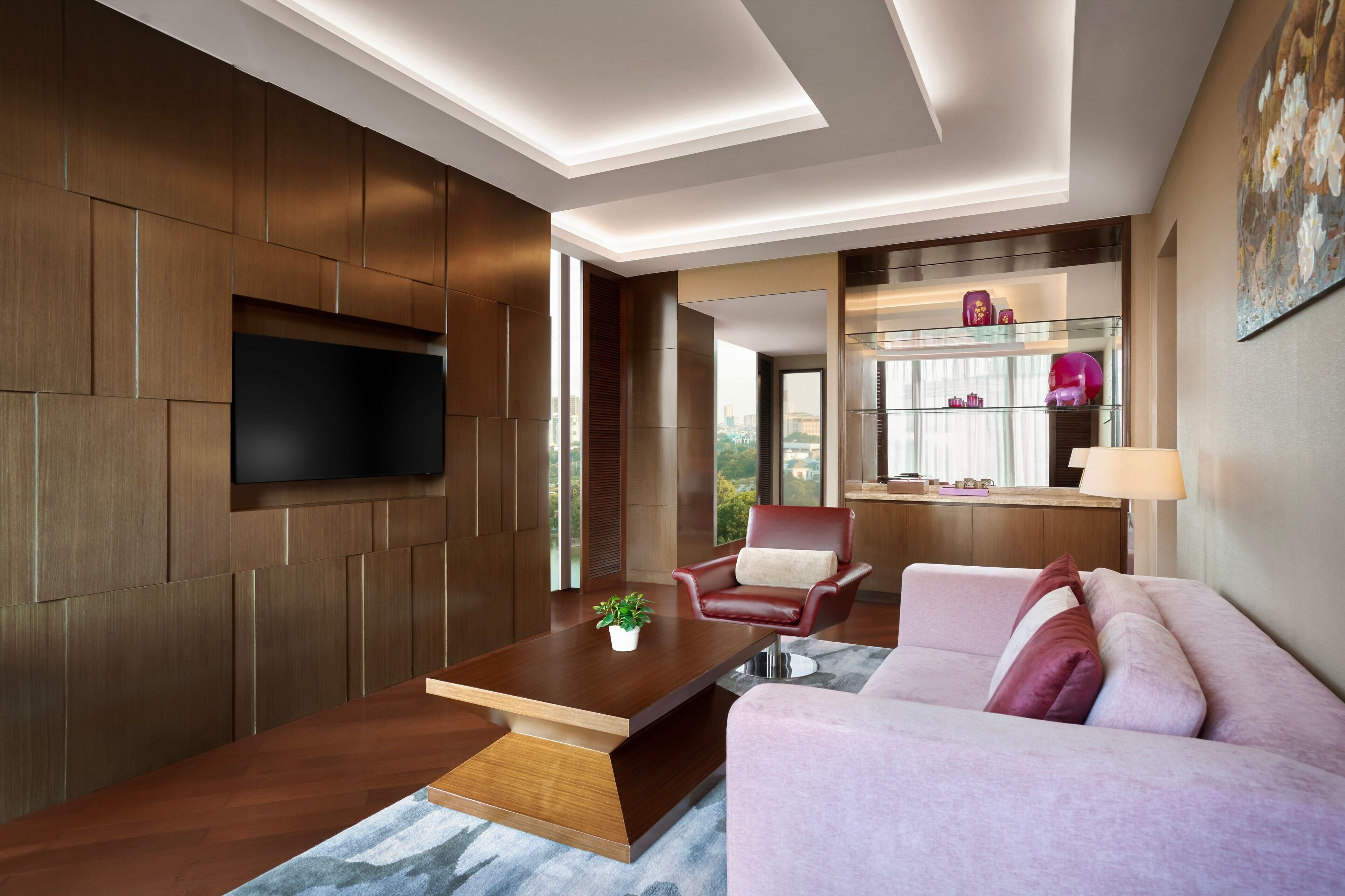 Una sala de estar moderna con pared de paneles de madera, televisor de pantalla plana, una mesita de centro con una planta, un sillón de cuero rojo, un sofá rosa y un aparador de vidrio con objetos decorativos.