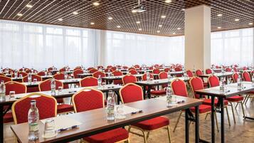 Un salón de conferencias con filas de mesas y sillas, ventanas amplias y un techo con diseño de cuadrícula.