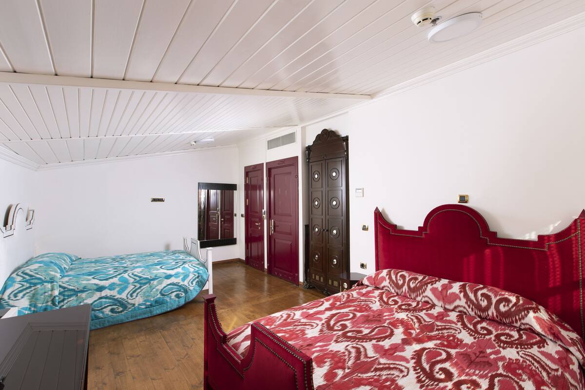 A bedroom with a wooden ceiling, two beds with patterned bedding, a wardrobe, and a mirror.