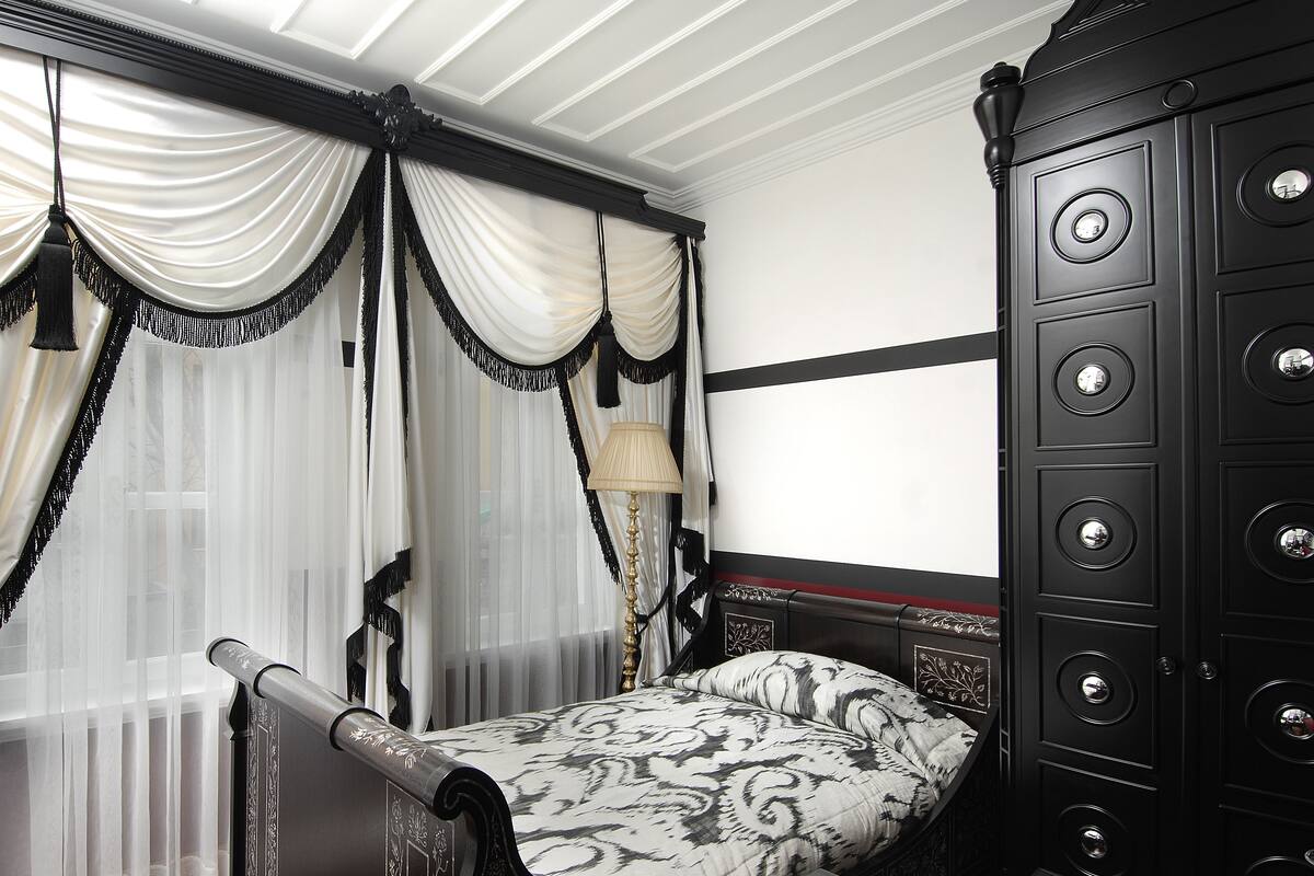 A bedroom with a bed, a nightstand, a lamp, a dresser, and a window with curtains.