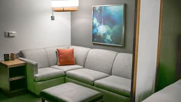 A modern hotel room with a grey sectional sofa, a small wooden nightstand, a green ottoman, a wall-mounted lamp, and a large abstract painting.