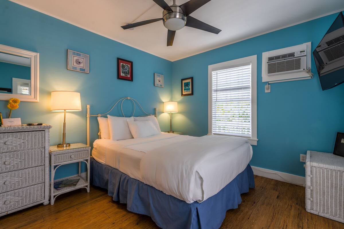 A bedroom with a bed, two bedside tables, a dresser, a ceiling fan, and wall-mounted air conditioning units.