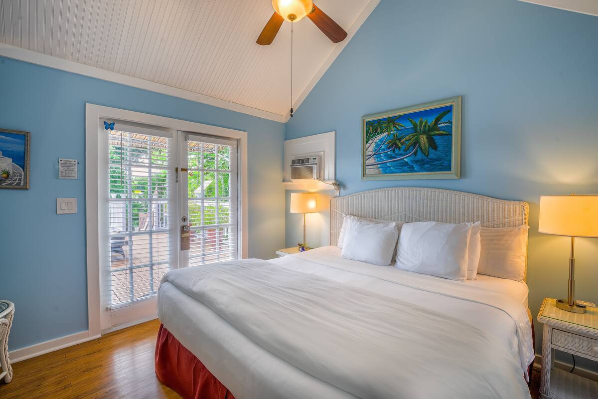 A bedroom with a bed, a ceiling fan, a window with blinds, a painting, and a nightstand with a lamp.