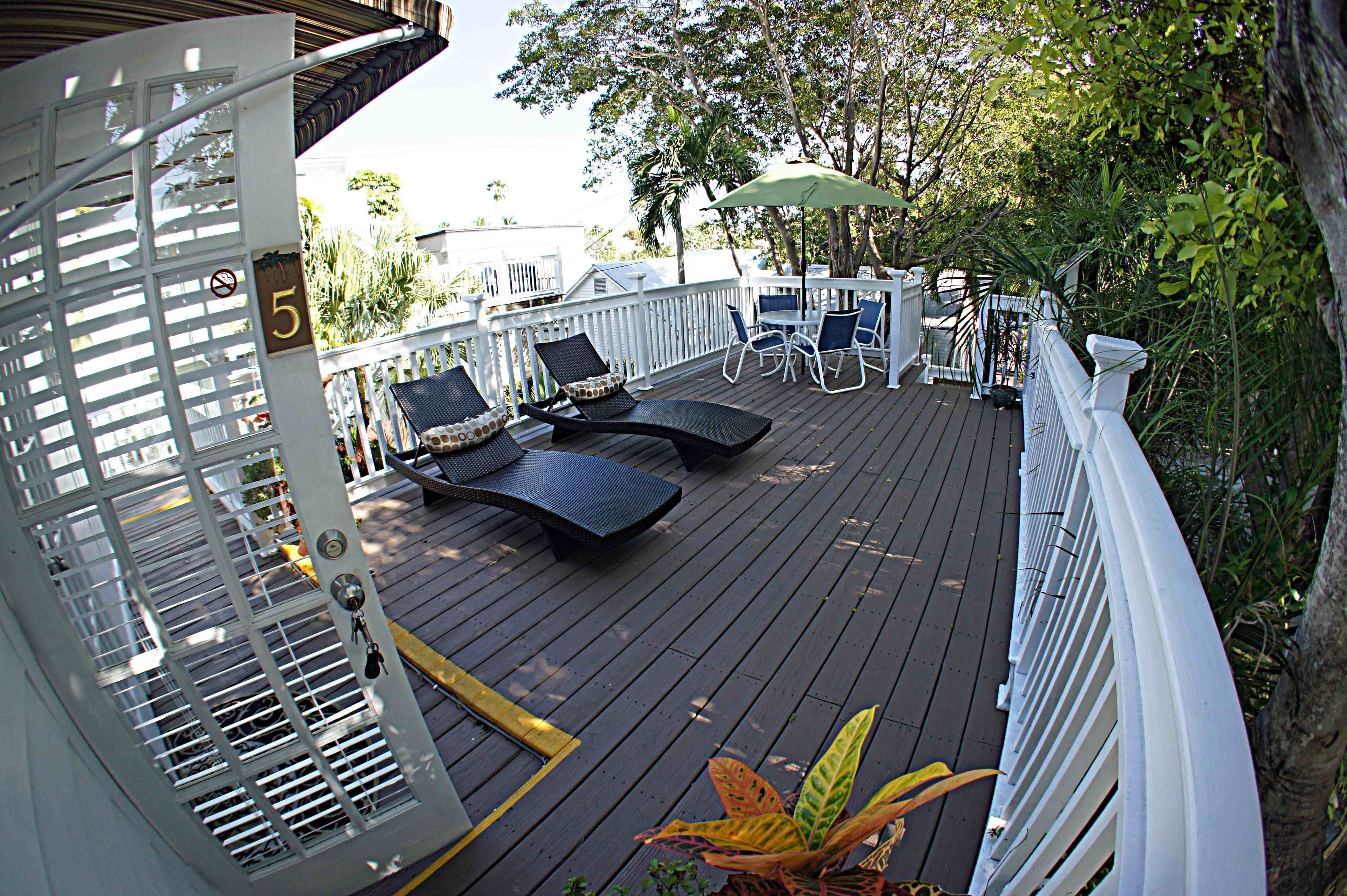 Eine Holzterrasse mit Liegestühlen und einem Tisch, umgeben von weißem Geländer und viel Grün.