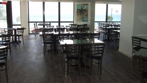 Ein Restaurant mit mehreren Tischen und Stühlen, großen Fenstern mit Meerblick und einem Palmenbild an der Wand.