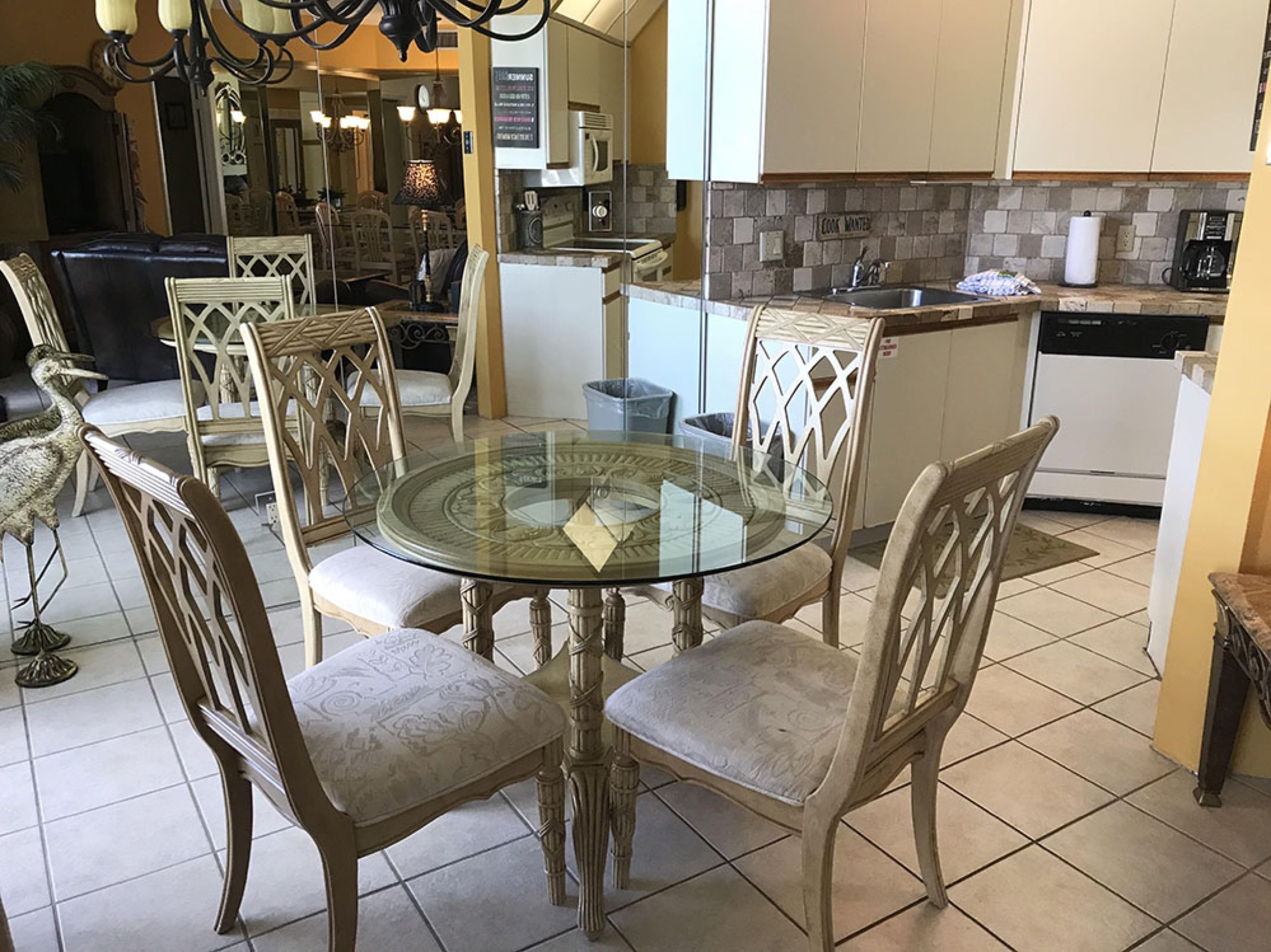 A dining area with a glass-top table surrounded by upholstered chairs, a kitchen with a stone backsplash, and a visible trash can.