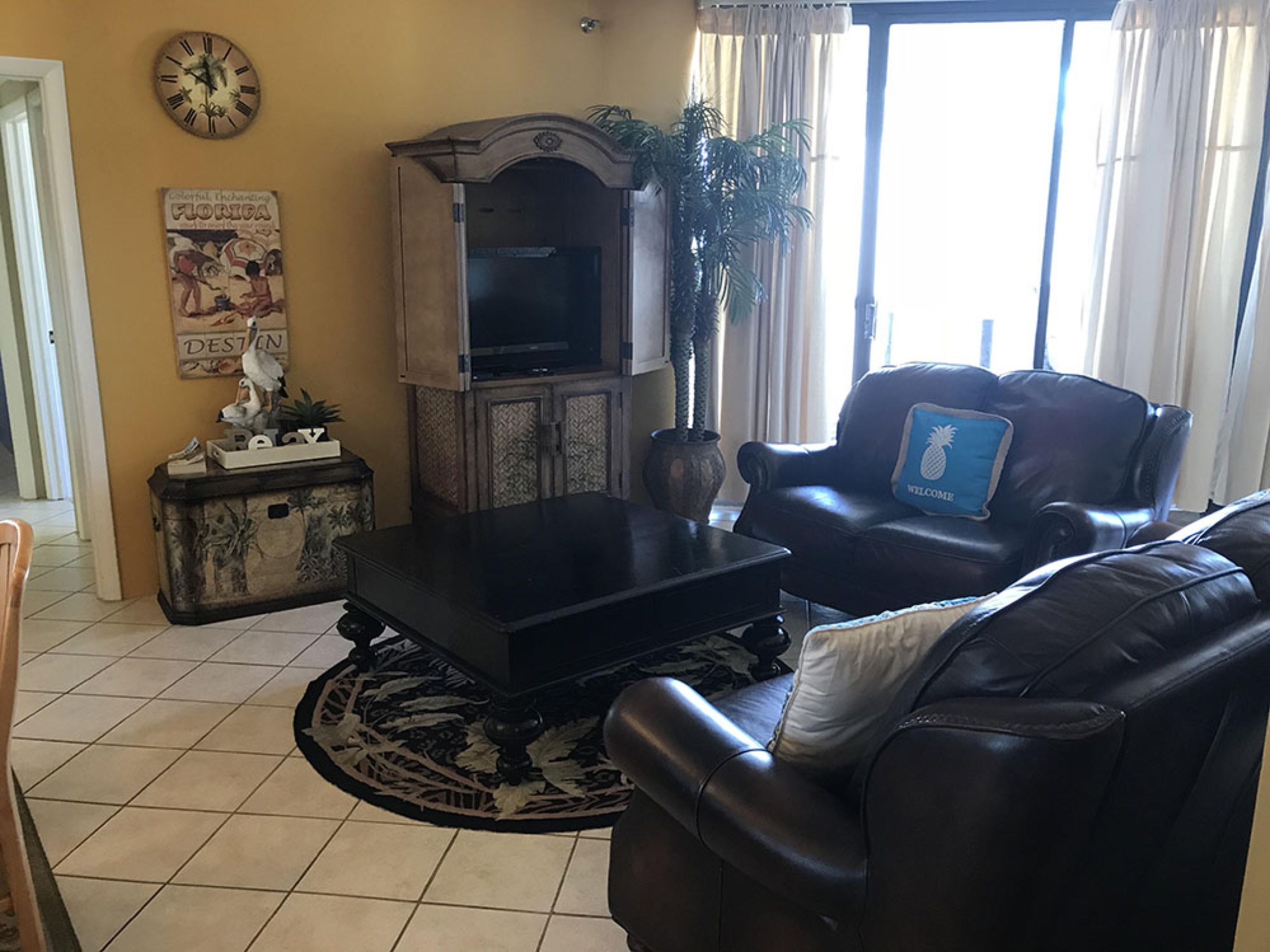 Un salon avec un canapé en cuir noir, une table basse, un téléviseur posé sur un meuble, une horloge au mur et une plante.
