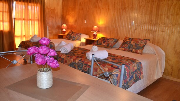A hotel room with two beds, a wooden wall, a table with a vase of flowers, and two bedside lamps. - Turismo Vinapu (Hanga Roa)