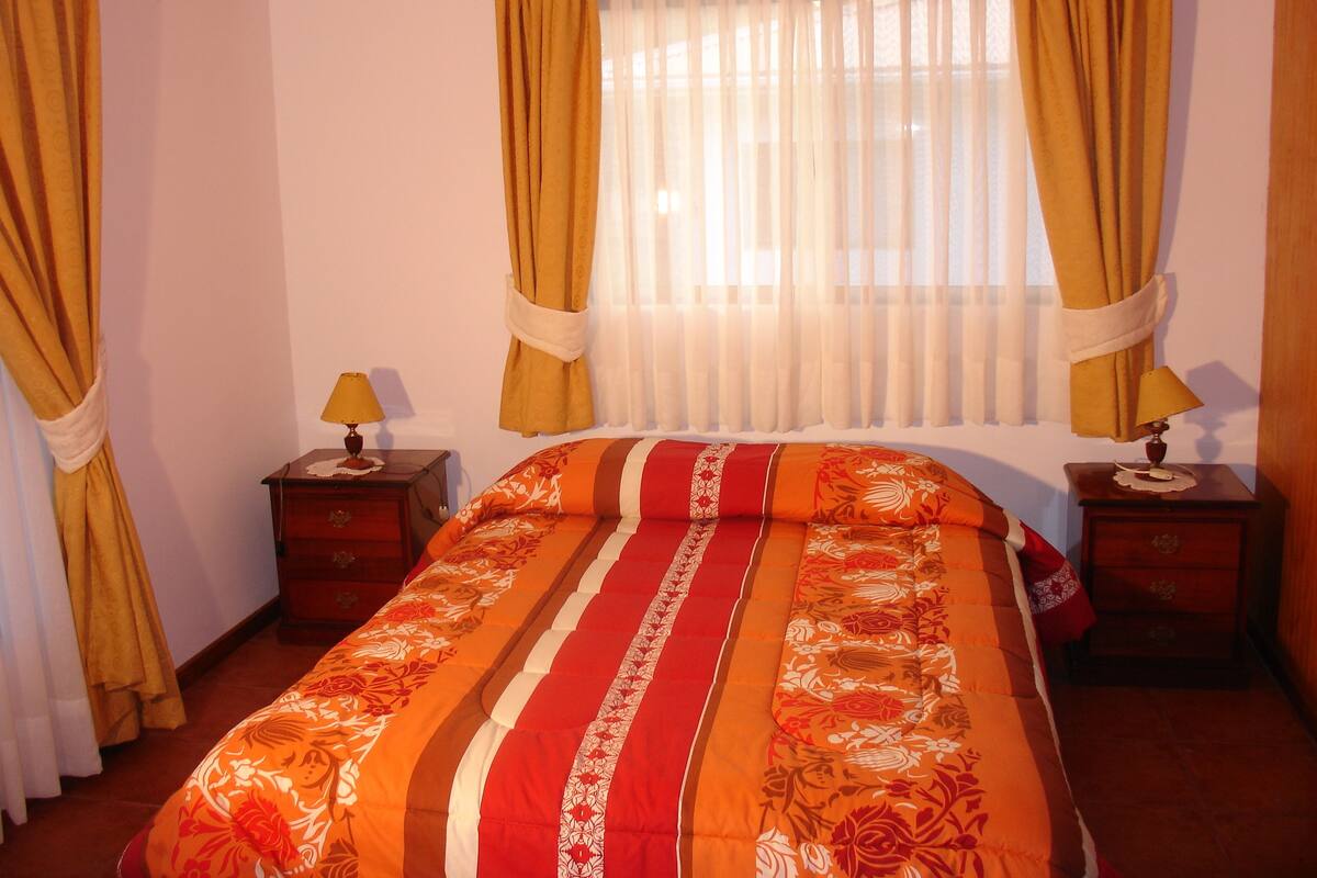 Un dormitorio con una cama, dos mesitas de noche, una ventana con cortinas y una lámpara en cada mesita.