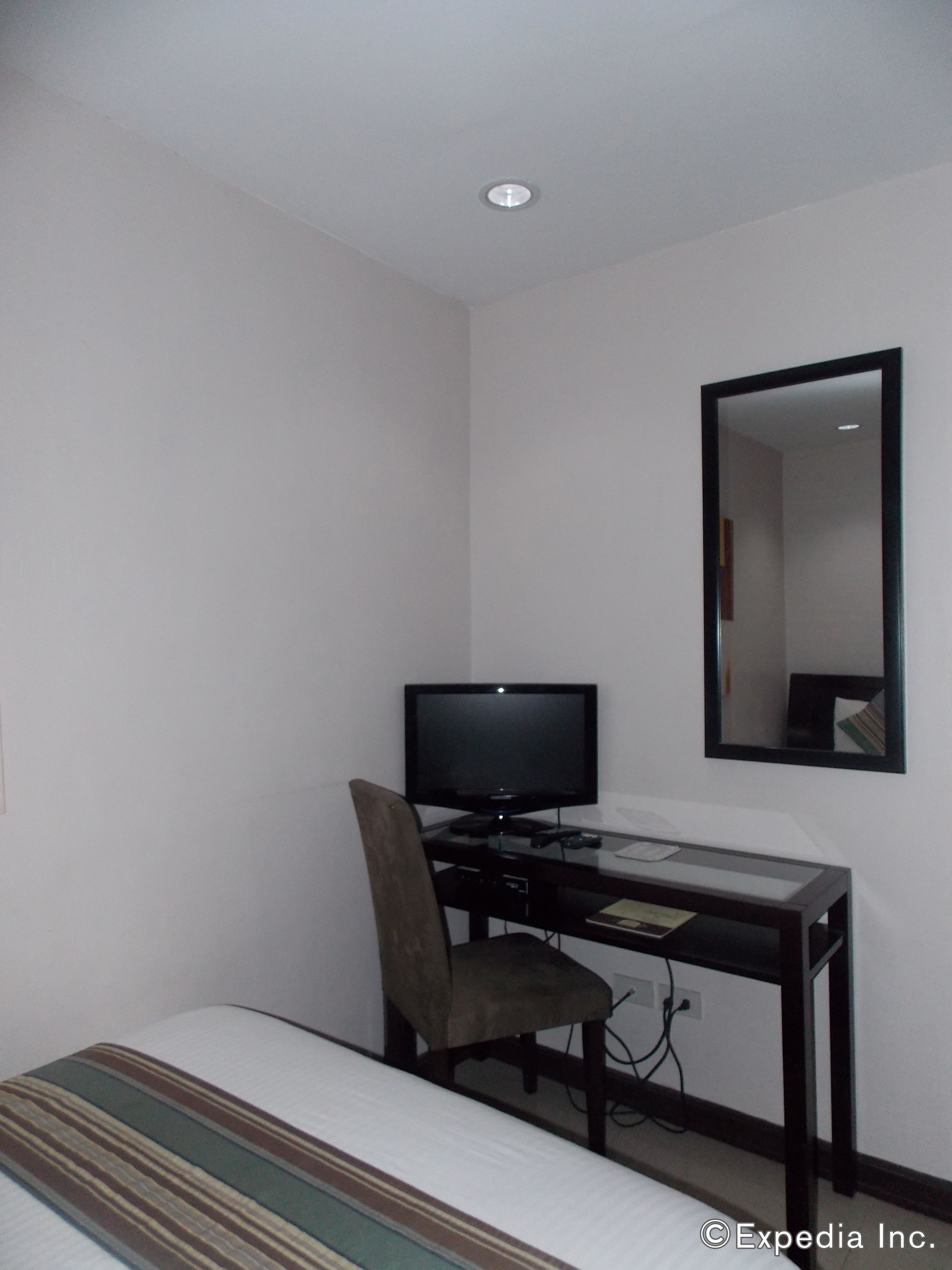 Een kleine, eenvoudige kamer met een bed, een bureau met een computer en monitor, en een spiegel aan de muur.