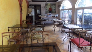 A dining area with multiple tables and chairs, a kitchen in the background, and large windows offering a view of the outdoors. - The Suites at Calle Nueva (Bacolod)