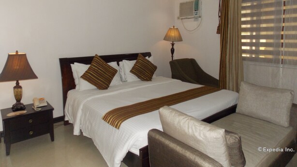 A hotel room with a bed, bedside tables, a lamp, a chair, a window with blinds, and an air conditioning unit. - The Suites at Calle Nueva (Bacolod)