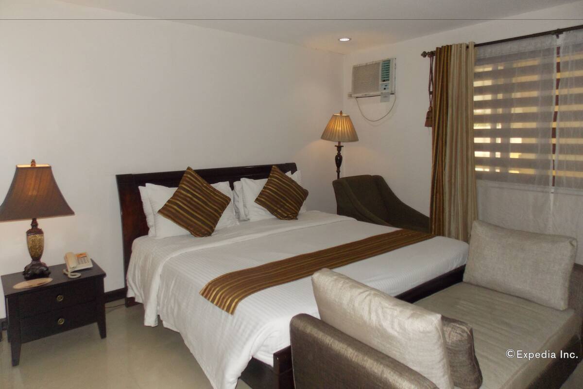 Une chambre d’hôtel comprenant un lit, des tables de chevet, une lampe, une chaise, une fenêtre avec des stores et un appareil de climatisation.