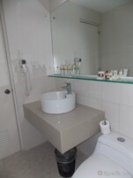Un baño con lavabo blanco, repisa de vidrio con artículos de aseo, secador de pelo y cubo de basura.