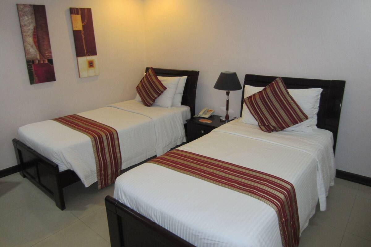 Une chambre d’hôtel avec deux lits simples, chacun recouvert d’une couvre-lit à rayures et agrémenté d’oreillers. Une table de chevet avec une lampe et un téléphone se trouve entre les deux lits.