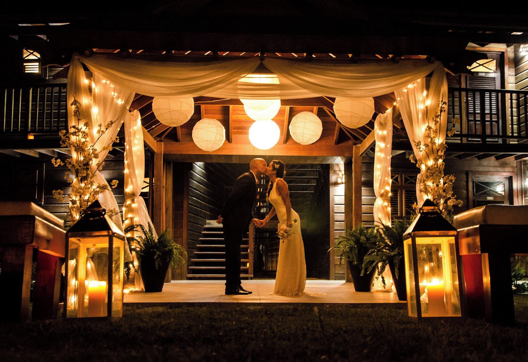 A couple kissing under a canopy with string lights and lanterns, with a staircase and wooden walls in the background.