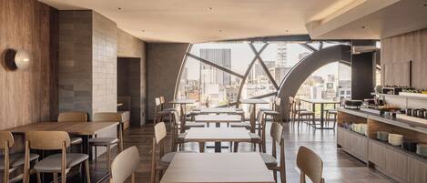 A modern dining area with tables and chairs, a kitchen counter, and a view of a cityscape through a unique architectural feature.