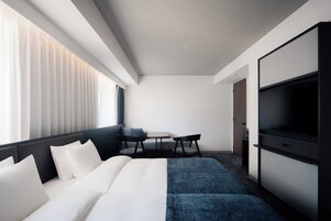 Una camera d'albergo moderna con un letto, un'area salotto con un tavolo, una TV e un'ampia finestra con persiane.