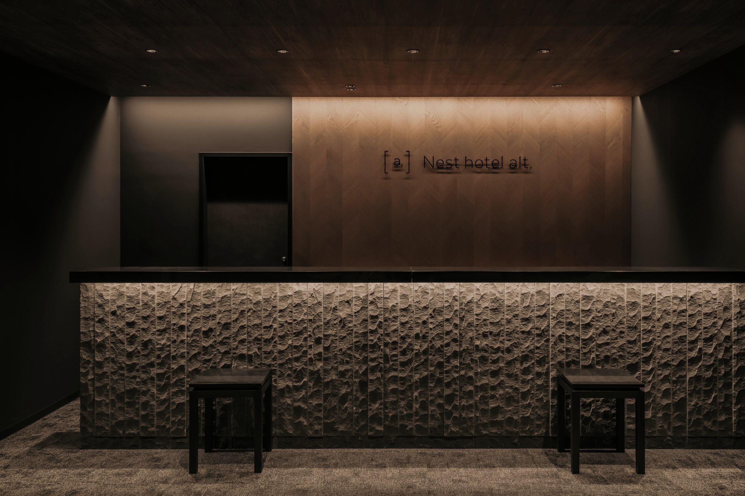 A reception desk with a wooden panel and a stone-textured counter. There are two stools and a door to the left.