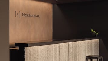 A reception desk with a wooden ceiling and a wall-mounted sign that reads 'Nest hotel alt.' There are two small tables in front of the desk.