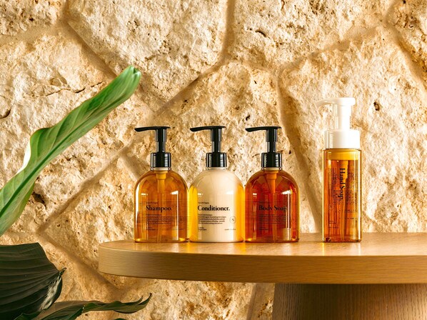 Une étagère en bois sur laquelle sont posées quatre bouteilles de produits d’hygiène personnelle, le tout adossé à un mur en pierre à la texture particulière.