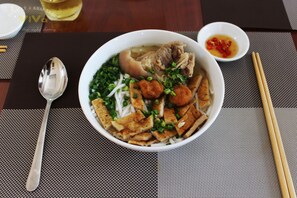 A bowl of noodle soup with tofu and vegetables, a spoon, chopsticks, and a small bowl of sauce on a table.