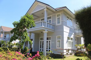 A two-story house with a balcony, surrounded by greenery and flowering plants.