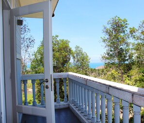 A balcony with white balustrades overlooking a lush garden and a distant view of the ocean.