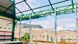Una terraza en la azotea con barandilla de vidrio, una zona de bar con taburetes y vistas a un paisaje urbano.