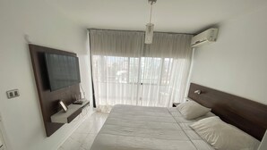A bedroom with a bed, a TV mounted on the wall, an air conditioning unit, and a window with sheer curtains.