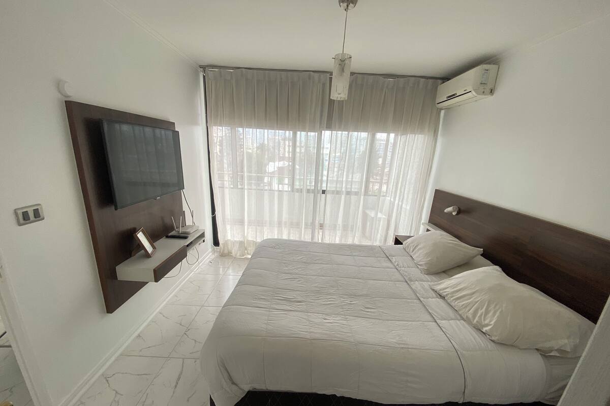 A bedroom with a bed, a TV mounted on the wall, an air conditioning unit, and a window with sheer curtains.